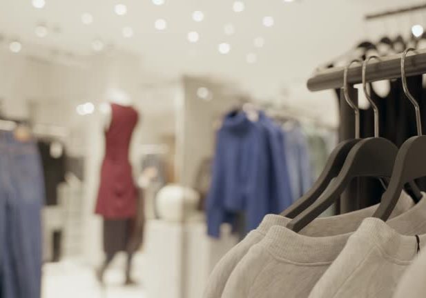 Clothes and wear items in display in shopping center or fashion store indoors. New clothes on shelves and rack hangers while shoppers walk around looking to buy latest fashion on sale in mall